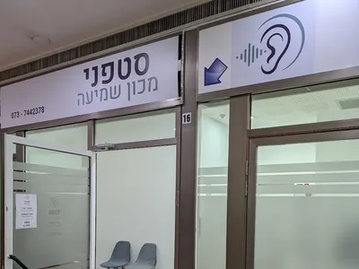 דלת הכניסה הזכוכית של המכון הפונה למסדרון, עם שלט המכון, סמל אוזן ושני כיסאות הנראים דרך הדלת הפתוחה.